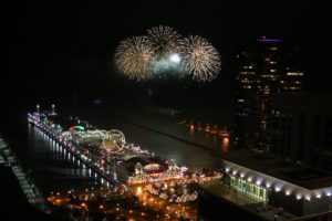 Fireworks light up the night sky over a brightly lit pier with a Ferris wheel and surrounding city buildings.