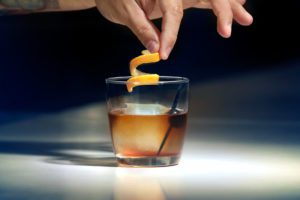 A hand garnishes a glass of whiskey on the rocks with orange peel—a classic Chicago touch perfect for your next entertainment or hotel Chicago stay.