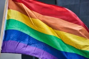 A rainbow-colored pride flag with six horizontal stripes waves in the wind against a gray background.